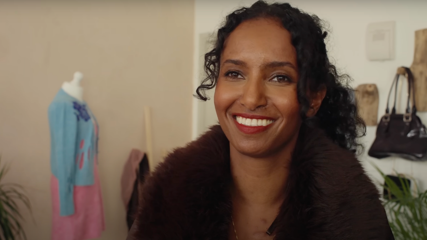 A smiling woman with dark curly hair wearing a brown fur-lined coat, sitting in a warmly lit fashion studio with a mannequin dressed in a blue and pink outfit in the background.